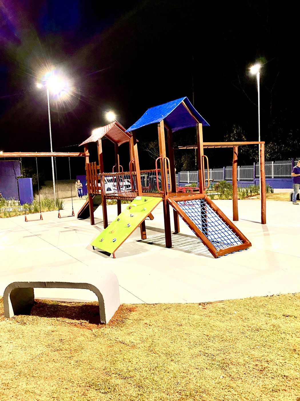 Playground de madeira de eucalipto em praça iluminada à noite, com rampa de rede e parede de escalada